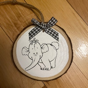 Puede incluir: Un adorno redondo de madera blanca con un lazo a cuadros blanco y negro. El adorno presenta una ilustración en blanco y negro de Dumbo el elefante.