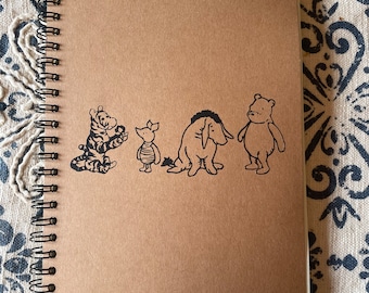 May include: A brown spiral bound notebook featuring a black and white illustration of Winnie the Pooh and his friends Piglet, Eeyore, and Tigger.