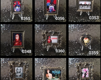 May include: A collection of miniature framed horror movie posters, each with a unique design and displayed on small clear stands. The frames are ornate and metallic, with the posters featuring various movie titles and imagery. The collection includes posters with the numbers 0365, 0356, 0353, 0348, 0360, 0359, 0339, 0341, and 0363.