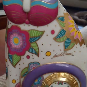 May include: A white ceramic cat figurine featuring a colorful floral pattern in pink, green, and blue, with gold accents. The cat has a purple curled tail that frames a round clock face with gold Roman numerals and a signature by Laurel Burch.