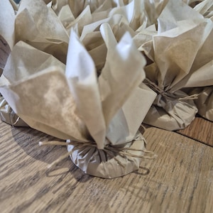 May include: A selection of small, round objects wrapped in brown paper and tied with twine. The paper is folded to create a decorative, flower-like appearance. The objects are arranged on a light brown wooden surface.