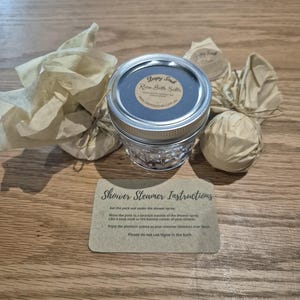 以下が含まれることがあります： ローズバスソルトが入ったガラス瓶。瓶には「Sleepy Song Rose Bath Salts」とウェブサイトのアドレスが書かれた茶色のラベルが貼られています。瓶の周りは、紐で結ばれた小さな白い紙袋が3つあります。手前に、バスソルトの使い方の説明が書かれた小さなカードがあります。カードには「Shower Steamer Instructions」と書いてあります。