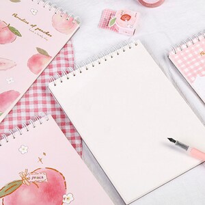K&ouml;nnte beinhalten: Spiralgebundene Notizb&uuml;cher mit Pfirsich-Illustrationen. Die Notizb&uuml;cher haben rosa Einb&auml;nde mit Pfirsich- und Blumendesigns. Ein Notizbuch ist mit einem Stift ge&ouml;ffnet, und der Text "Paradise of peaches" ist auf dem Einband zu sehen.