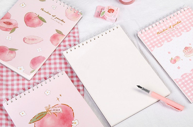 K&ouml;nnte beinhalten: Spiralgebundene Notizb&uuml;cher mit Pfirsich-Illustrationen. Die Notizb&uuml;cher haben rosa Einb&auml;nde mit Pfirsich- und Blumendesigns. Ein Notizbuch ist mit einem Stift ge&ouml;ffnet, und der Text "Paradise of peaches" ist auf dem Einband zu sehen.