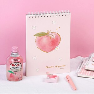 K&ouml;nnte beinhalten: Ein rosa Spiralnotizblock mit einer Pfirsichillustration, eine Flasche Pfirsichgetr&auml;nk, eine kleine Schachtel mit Pfirsichthema und ein rosa Stift. Das Notizbuch tr&auml;gt die Worte "Paradise of peaches" und "Have a lovely day."
