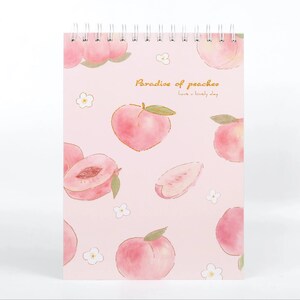 K&ouml;nnte beinhalten: Ein spiralgebundenes Notizbuch mit rosa Einband, verziert mit Aquarell-Illustrationen von Pfirsichen und kleinen wei&szlig;en Bl&uuml;ten. Auf dem Cover steht der Text "Paradise of peaches" und "have a lovely day".