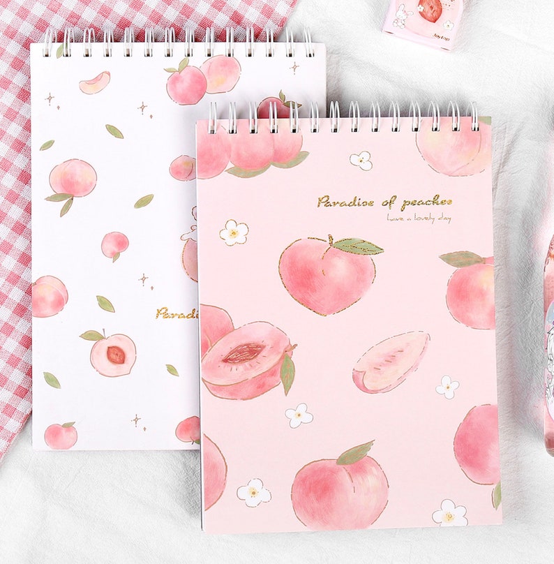 K&ouml;nnte beinhalten: Zwei Spiralnotizb&uuml;cher mit Pfirsichillustrationen. Eines hat einen wei&szlig;en Hintergrund mit Pfirsich- und Blattmustern. Das andere hat einen rosa Hintergrund mit Pfirsichillustrationen und dem Text "Paradise of peaches".