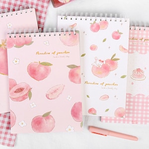 K&ouml;nnte beinhalten: Vier Spiralnotizb&uuml;cher mit Pfirsichmotiven. Die Notizb&uuml;cher zeigen Aquarellillustrationen von Pfirsichen, Blumen und einem Hasen. Der Text "Paradise of peaches" ist auf einigen Umschl&auml;gen aufgedruckt. Ein rosa Stift liegt auf der Oberfl&auml;che.
