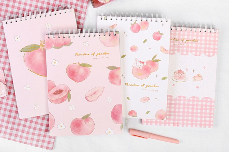 K&ouml;nnte beinhalten: Vier Spiralnotizb&uuml;cher mit Pfirsichmotiven. Die Notizb&uuml;cher zeigen Aquarellillustrationen von Pfirsichen, Blumen und einem Hasen. Der Text "Paradise of peaches" ist auf einigen Umschl&auml;gen aufgedruckt. Ein rosa Stift liegt auf der Oberfl&auml;che.