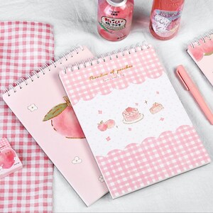 K&ouml;nnte beinhalten: Rosa Spiralbl&ouml;cke mit Pfirsich-Illustrationen und dem Text "Paradise of peaches". Ein Notizblock zeigt eine Pfirsich-Grafik, ein anderer ein rosa Vichy-Muster und Kuchen-Illustrationen. Ein rosa Stift und rosa karierter Stoff sind ebenfalls zu sehen.