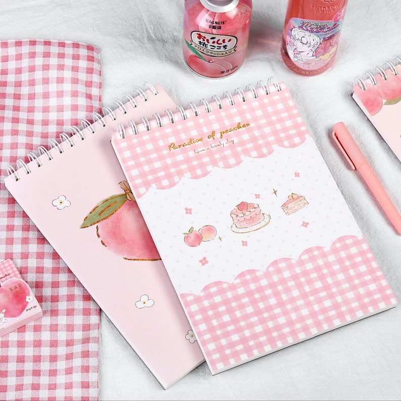 K&ouml;nnte beinhalten: Rosa Spiralbl&ouml;cke mit Pfirsich-Illustrationen und dem Text "Paradise of peaches". Ein Notizblock zeigt eine Pfirsich-Grafik, ein anderer ein rosa Vichy-Muster und Kuchen-Illustrationen. Ein rosa Stift und rosa karierter Stoff sind ebenfalls zu sehen.
