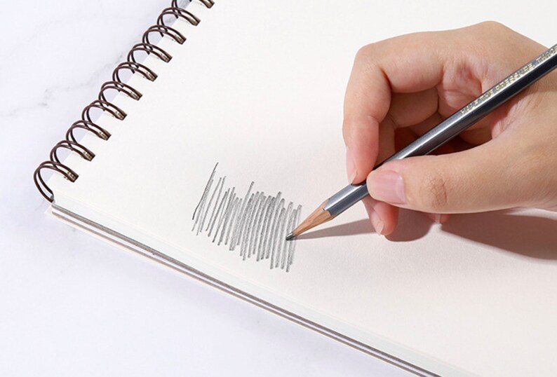 K&ouml;nnte beinhalten: Eine Hand, die einen grauen Bleistift h&auml;lt und auf einem wei&szlig;en Spiralnotizblock skizziert. Der Bleistift erzeugt eine Reihe grauer Linien auf dem Papier, was die Verwendung des Bleistifts zum Zeichnen oder Schreiben demonstriert. Die Spiralbindung des Notizblocks ist sichtbar.