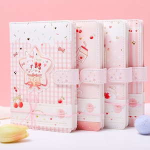 May include: A set of four pink and white notebooks with cute illustrations. The notebooks feature a star-shaped design with a bunny and cupcake, cherries, and the text "sweet so". Each notebook has a pink strap closure.