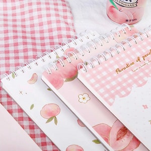 K&ouml;nnte beinhalten: Eine Sammlung von Spiralnotizb&uuml;chern mit Pfirsichmotiven. Die Notizb&uuml;cher zeigen Illustrationen von Pfirsichen, Blumenmustern und den Text "Paradise of peaches". Die Farbpalette umfasst rosa, wei&szlig;e und goldene Akzente.