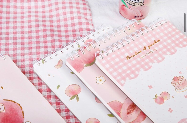 K&ouml;nnte beinhalten: Eine Sammlung von Spiralnotizb&uuml;chern mit Pfirsichmotiven. Die Notizb&uuml;cher zeigen Illustrationen von Pfirsichen, Blumenmustern und den Text "Paradise of peaches". Die Farbpalette umfasst rosa, wei&szlig;e und goldene Akzente.