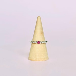 May include: A silver beaded ring with a central magenta bead is displayed on a wooden cone-shaped stand. The ring features small, round silver beads. The background is a soft pink.