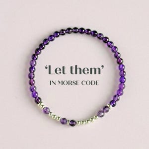 May include: A beaded bracelet with purple amethyst stones and silver beads. The bracelet is circular and features the text "Let them" in Morse code. The background is a soft pink.