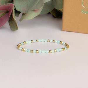 May include: An elegant bracelet with light blue beads and gold-coloured beads. The bracelet is presented on a pale pink surface, with a gold-coloured box in the background. A stylish piece of jewellery.