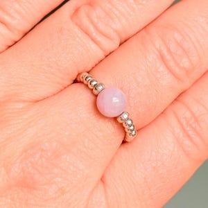 May include: A silver beaded ring with a light pink, round gemstone at the center. The ring is on a finger, showcasing the delicate design and color contrast. The beads are small and evenly spaced.