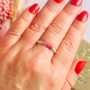 May include: A silver ring with a row of small, faceted pink and red gemstones. The ring is on a person's finger with red nail polish. The ring is a delicate piece of jewelry.