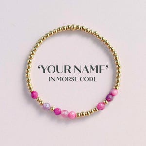 May include: Gold beaded bracelet with pink and purple gemstone accents. The bracelet features the text "YOUR NAME" in Morse code. The beads are arranged in a circular pattern against a light pink background.