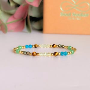 May include: A colourful beaded bracelet with green, blue, yellow, and brown gemstones on a gold-coloured chain. The bracelet is displayed on a light pink surface, with a blurred background. The bracelet is approximately 18 cm in length.