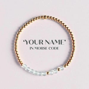 May include: Gold beaded bracelet featuring clear crystal beads. The text "YOUR NAME" in Morse code is displayed on a pink background. A stylish piece of jewellery.
