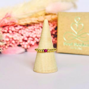 May include: A gold beaded ring with a central red gemstone is displayed on a wooden cone. The ring is set against a backdrop of pink flowers and a gold box with the text "Soul Seekers".