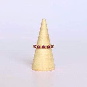 May include: A gold ring featuring alternating small, round, ruby-coloured beads and gold beads. The ring is displayed on a light-coloured wooden cone-shaped ring holder against a white background. The ring is a delicate piece of jewellery.