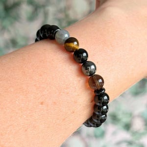 May include: A beaded bracelet featuring a mix of black, gray, brown, and gold-colored gemstones. The bracelet is strung on an elastic band and is worn on a wrist. The gemstones are round and polished.