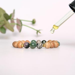 May include: A beaded bracelet featuring a mix of wooden and gemstone beads in shades of brown, green, purple, and gray. The bracelet is accented with gold-colored beads. A dropper dispenses a clear liquid.