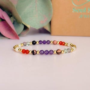 May include: A beaded bracelet featuring a variety of colorful gemstones and gold-colored beads. The gemstones include red, purple, pink, green, and black beads. The bracelet is displayed on a white surface with a blurred background.