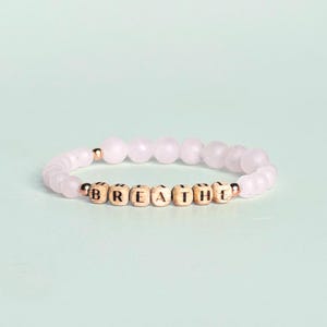 May include: A beaded bracelet with the word "BREATHE" spelled out in wooden letter beads. The bracelet features light pink, round gemstone beads and gold-colored accents. The background is a soft, light green.