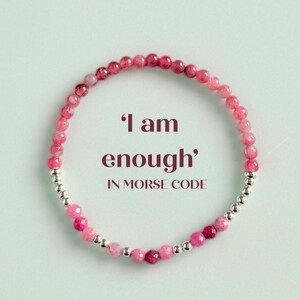 May include: A pink beaded bracelet with the words "I am enough" in Morse code. The bracelet features small, round, pink beads and silver-coloured spacer beads. The background is a light teal colour.