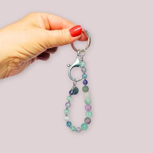 Könnte beinhalten: Ein Schlüsselanhänger mit Perlen, einem silbernen Karabiner und einem Schlüsselring. Die Perlen sind eine Mischung aus Pastellfarben, darunter Grün, Lila und Blau. Der Schlüsselanhänger wird von einer Hand mit rotem Nagellack gehalten.