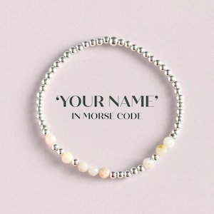 May include: A silver beaded bracelet with a mix of silver and light pink beads. The bracelet is circular and features the text "YOUR NAME" IN MORSE CODE. The background is a soft pink.