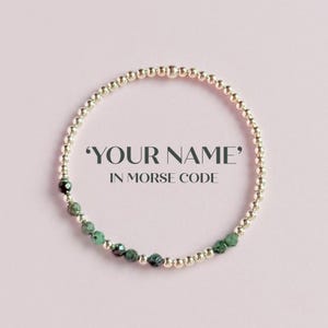 Könnte beinhalten: Ein silbernes Perlenarmband mit einem Abschnitt aus grünen Perlen. Das Armband wird auf einem rosa Hintergrund präsentiert. Der Text auf dem Bild lautet: "'YOUR NAME' IN MORSE CODE."