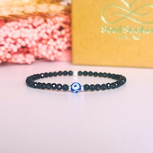 May include: Black beaded bracelet with a blue and white evil eye charm. The bracelet is made of small, faceted black beads and a central charm with a blue eye design. The background includes a gold box with the text "Soul Seeker Healing".