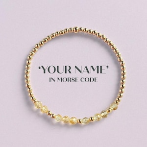 May include: A gold beaded bracelet with yellow gemstone accents. The bracelet is circular and features the text "YOUR NAME" in Morse code. The background is a soft purple.