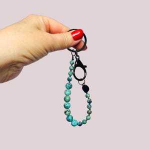 May include: A beaded keychain with a black clasp and ring. The beads are a mix of turquoise and green, varying in size. The keychain is held against a light pink background.