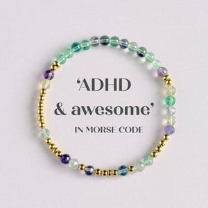 May include: A beaded bracelet with the text 'ADHD & awesome' in Morse code. The bracelet features a mix of colorful gemstones and gold-colored beads. The gemstones are in shades of green, purple, and blue.