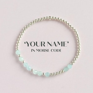 May include: A silver beaded bracelet with pale blue gemstone beads. The bracelet features the text 'YOUR NAME' IN MORSE CODE. The bracelet is set against a pale pink backdrop.
