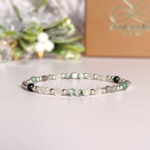 Könnte beinhalten: Ein Perlenarmband mit einer Mischung aus weißen, grünen und grauen Edelsteinen. Das Armband hat kleine, runde Perlen und silberfarbene Abstandshalter. Die Edelsteine variieren in Farbe und Textur und sind ein einzigartiges Accessoire.