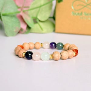 May include: A bracelet with wooden beads, gold spacers, and a selection of gemstones in various colours. The gemstones include purple, green, pink, and white. The bracelet is presented on a white surface.