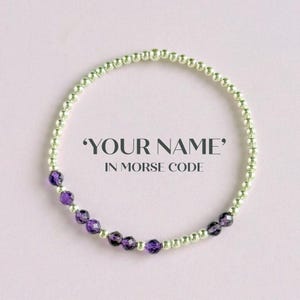 Könnte beinhalten: Ein silbernes Perlenarmband mit einem Abschnitt aus facettierten lila Perlen. Das Armband wird auf einem hellrosa Hintergrund präsentiert. Der Text auf dem Bild lautet 'YOUR NAME' IN MORSE CODE.