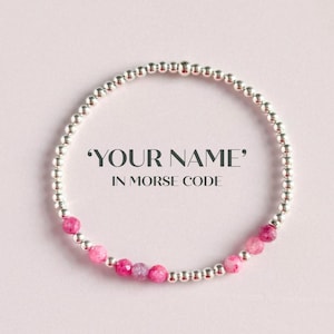 May include: A beaded bracelet with the text "YOUR NAME" in Morse code. The bracelet features small, silver-colored beads and larger, pink gemstone beads. The background is a soft pink.