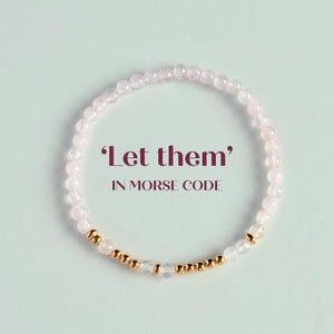 Könnte beinhalten: Ein zartes Armband aus kleinen, hellrosa Perlen und goldfarbenen Akzenten. Das Armband zeigt den Text „Let them“ in Morsecode, was ihm eine einzigartige Note verleiht. Das Design ist schlicht und elegant, geeignet für den täglichen Gebrauch.