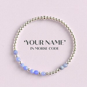 May include: A silver beaded bracelet with blue gemstone accents. The bracelet is circular, with the text "YOUR NAME" in Morse code. The background is a soft pink.