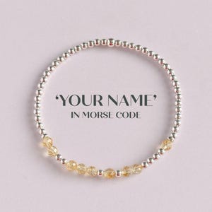 May include: A silver beaded bracelet with a section of yellow-gold gemstone beads. The bracelet is circular and features the text "YOUR NAME" in Morse code. The background is a soft, light purple.