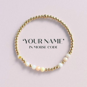 May include: Gold beaded bracelet with a Morse code design. The bracelet features a mix of gold and white beads, with the text "YOUR NAME" IN MORSE CODE. The bracelet is displayed on a light pink background.